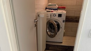 A compact laundry area with a front-loading washing machine, a drying rack, and a stack of folded towels. - 3-room vacation apartment, 64 square meters, with private parking in Ötlingen 79576, Weil am Rhein, near Basel/A5; accommodates up to 6 people (Weil am Rhein)