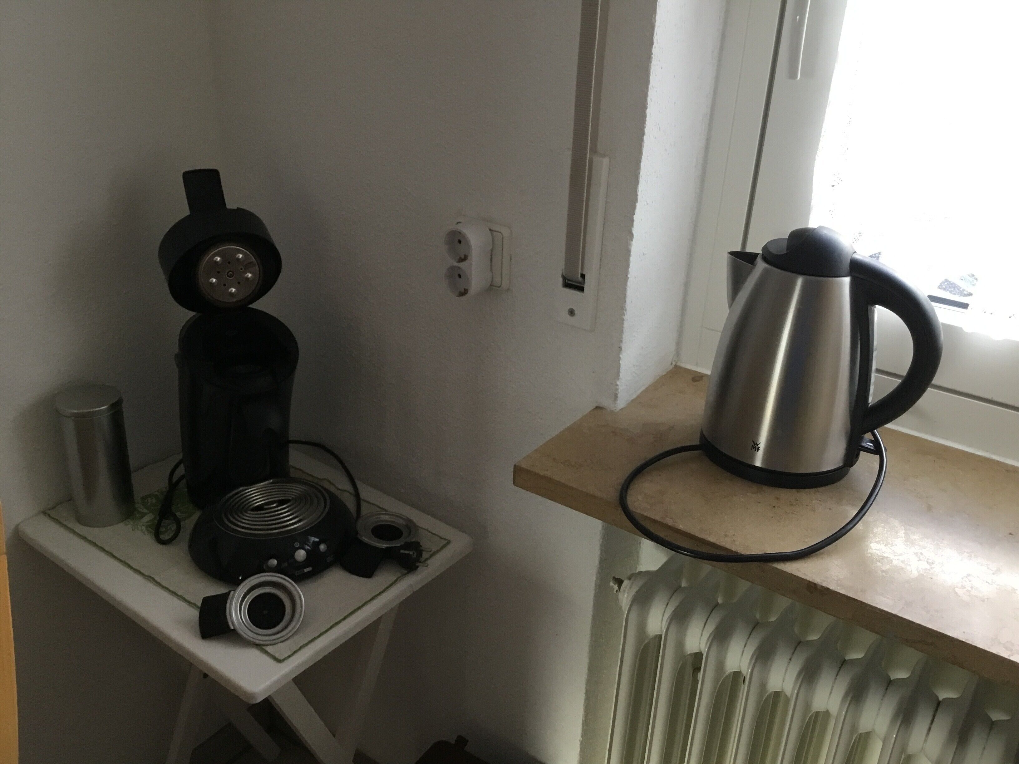 Cozinha compacta com cafeteira, chaleira e uma pequena mesa com um alto-falante e outros dispositivos eletrônicos.