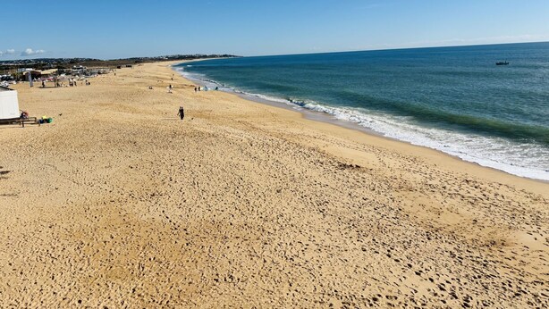 Een breed zandstrand met voetafdrukken, een heldere blauwe lucht en een rustige zee.