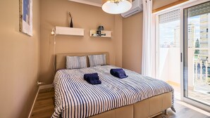 Een slaapkamer met een bed, nachtkastjes, een hanglamp en uitzicht op gebouwen door een raam.