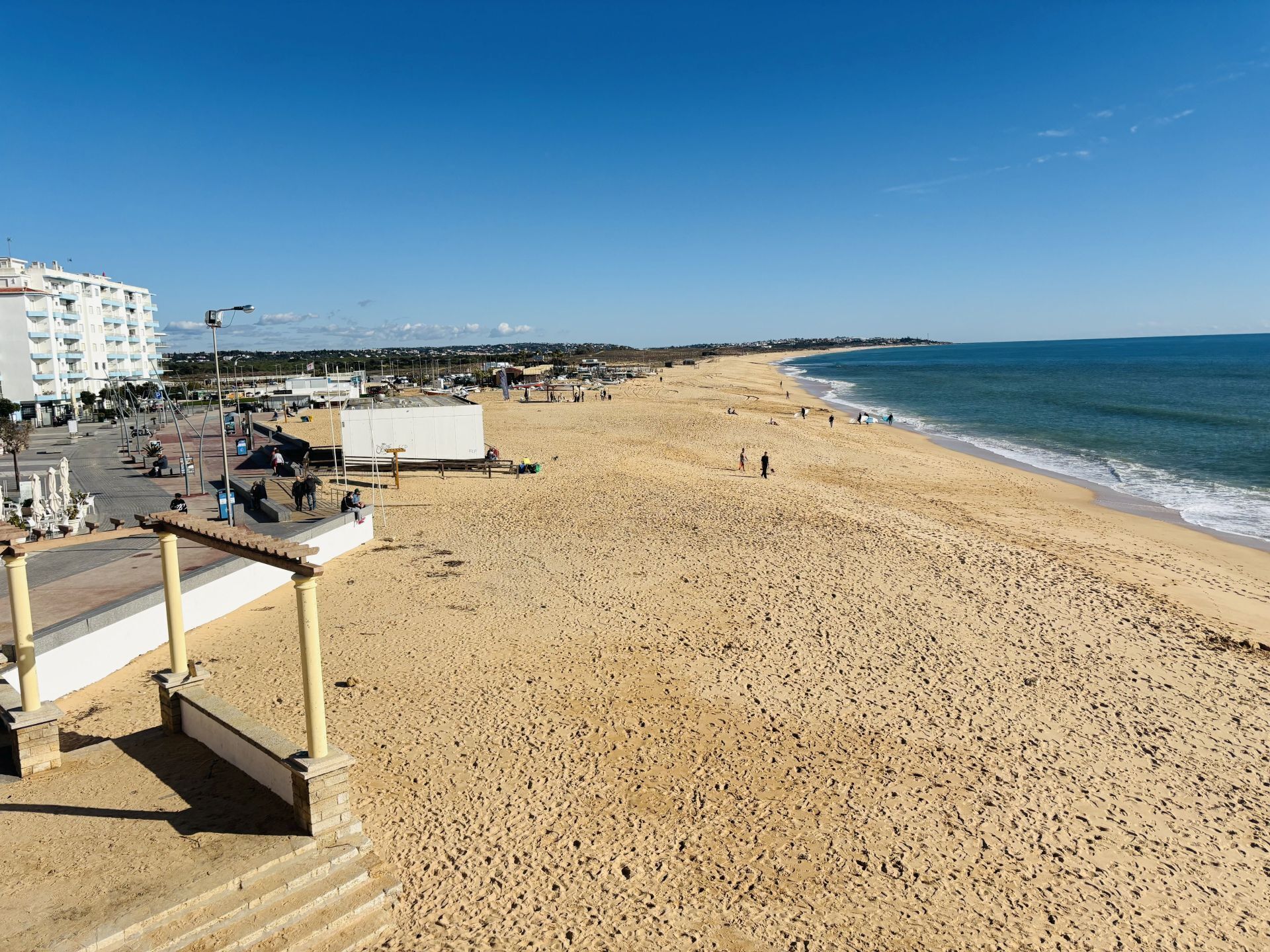 Une vaste plage de sable fin, un ciel bleu dégagé, quelques personnes et des bâtiments en arrière-plan.