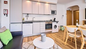 Een moderne keuken met witte kastjes, een magnetron, een fornuis en een wasmachine. Er is een eethoek met een tafel en stoelen, en een woonkamer met een bank.