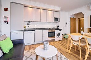 Une cuisine moderne avec des meubles blancs, un four à micro-ondes, une cuisinière et un lave-linge. On y trouve un coin repas avec une table et des chaises, ainsi qu’un salon avec un canapé.