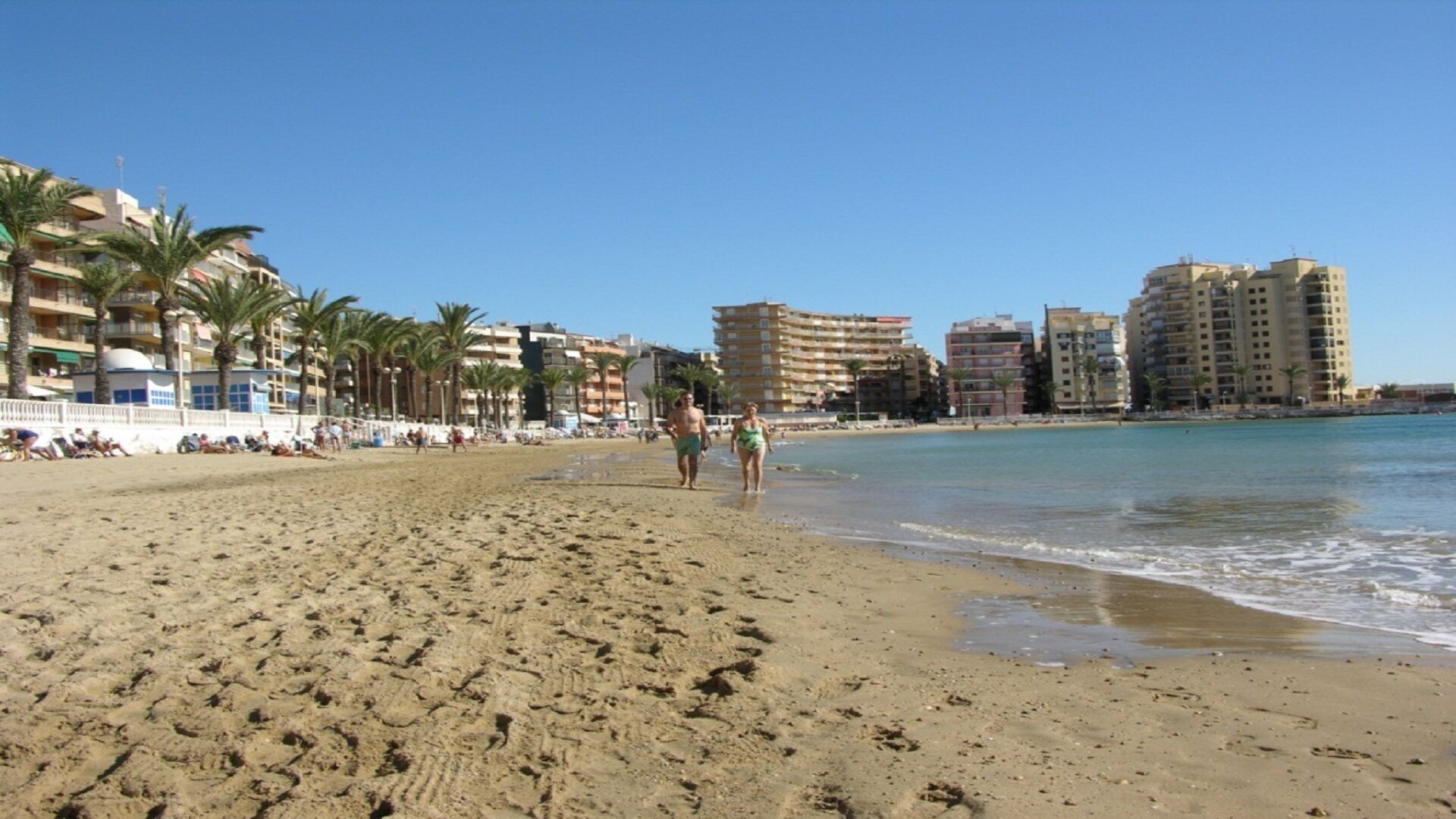A sandy beach with palm trees and a clear blue sky. There are buildings in the background, and people walking along the shore.