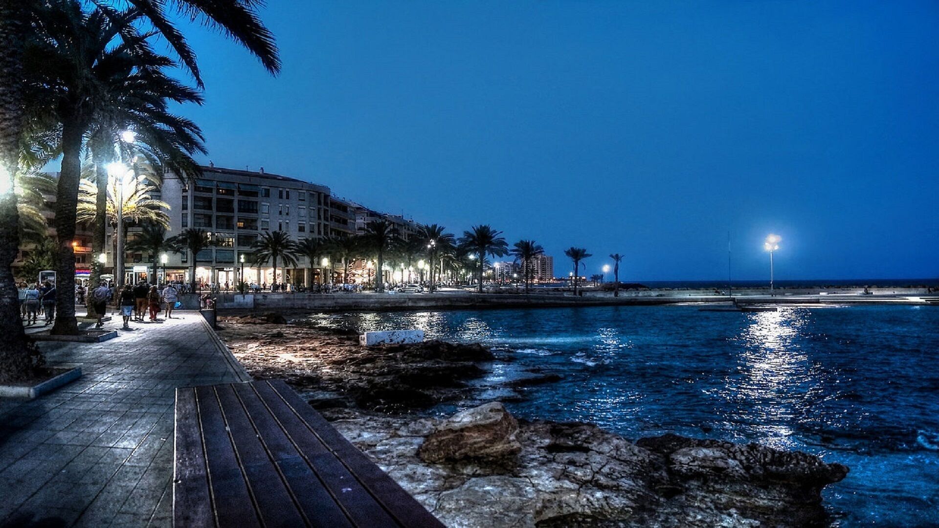 Un paseo marítimo con una pasarela de madera, palmeras y edificios al fondo.