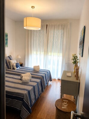 Ein Hotelzimmer mit Bett, Nachttisch, Lampe und Blick ins Freie durch durchsichtige Vorhänge.