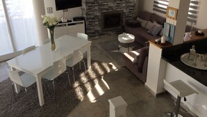 Un salon moderne avec une cheminée en pierre, un téléviseur à écran plat fixé au mur, un coin repas avec une table et des chaises blanches, et un espace salon confortable avec un canapé et une table basse.