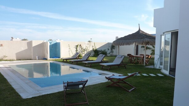 Un espace piscine avec des chaises longues, une structure à toit de chaume et un bâtiment blanc doté d’une porte en verre.