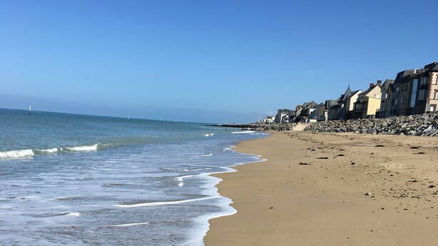 Une plage de sable où les vagues viennent lécher le rivage, une digue rocheuse et des bâtiments qui bordent la côte.