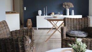 Un coin repas avec des chaises en osier, une table blanche et un mur foncé.