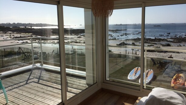 Una habitación con grandes ventanales que dan a una playa, una terraza de madera y vista al océano.