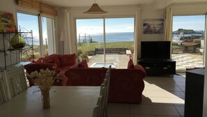 Amplia sala de estar con un gran ventanal con vistas a un paisaje costero, un sofá rojo confortable, una mesa de comedor con un jarrón de flores secas y un televisor de pantalla plana.