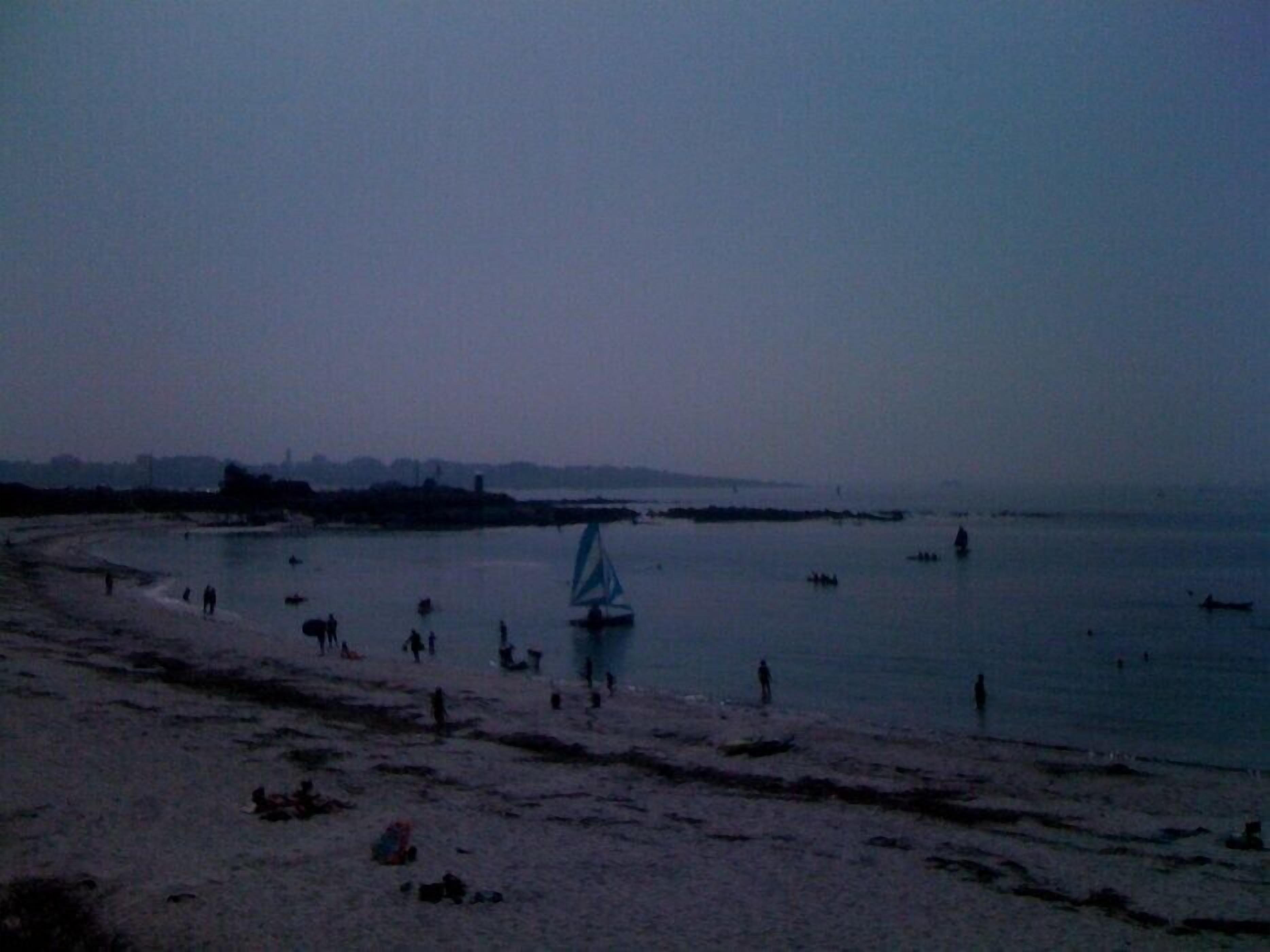 Une plage avec un voilier, des gens et, au loin, la silhouette d’une ville.
