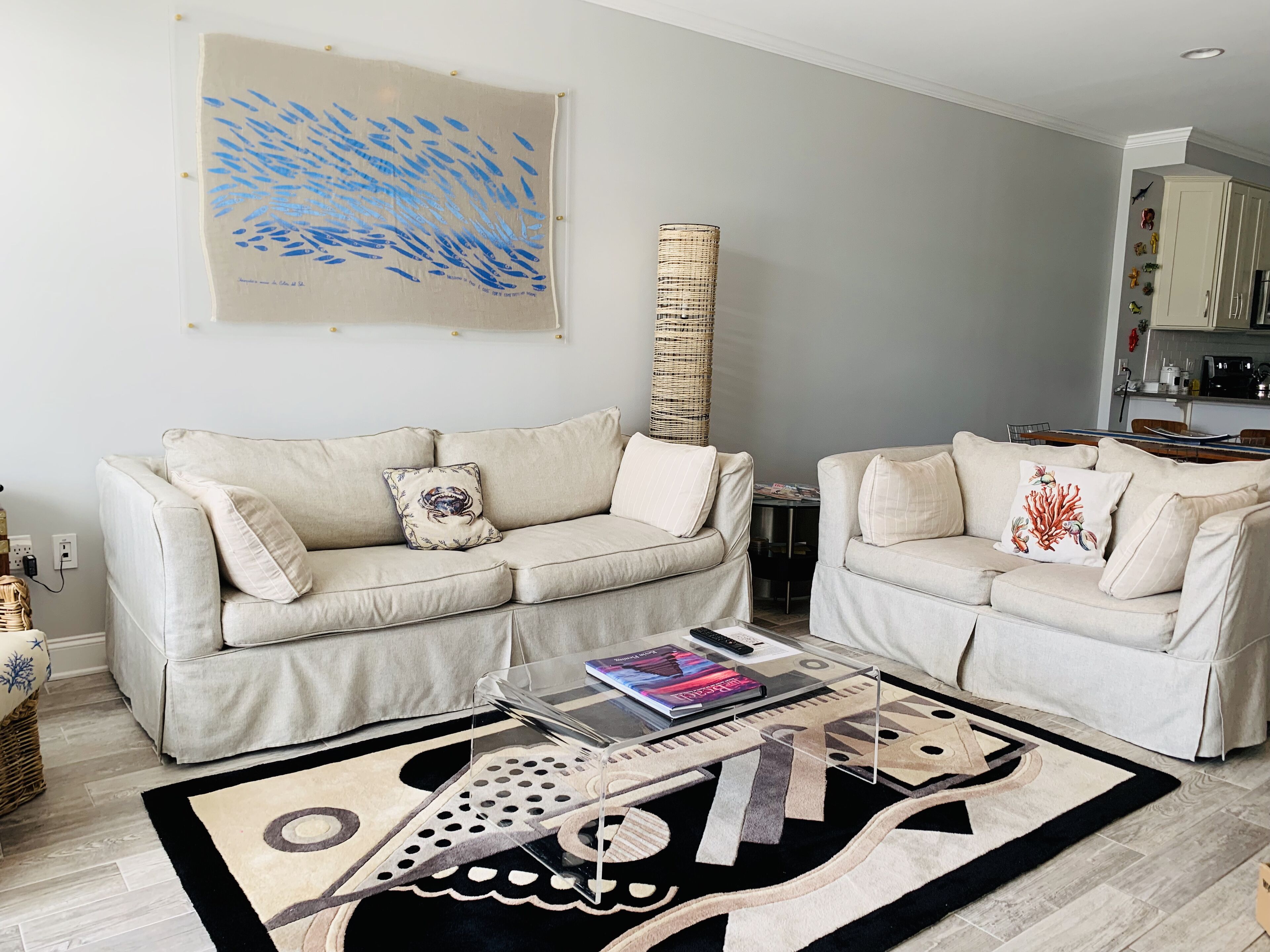 A living room with two beige sofas, a coffee table with magazines, a rug, and a wall-mounted artwork.
