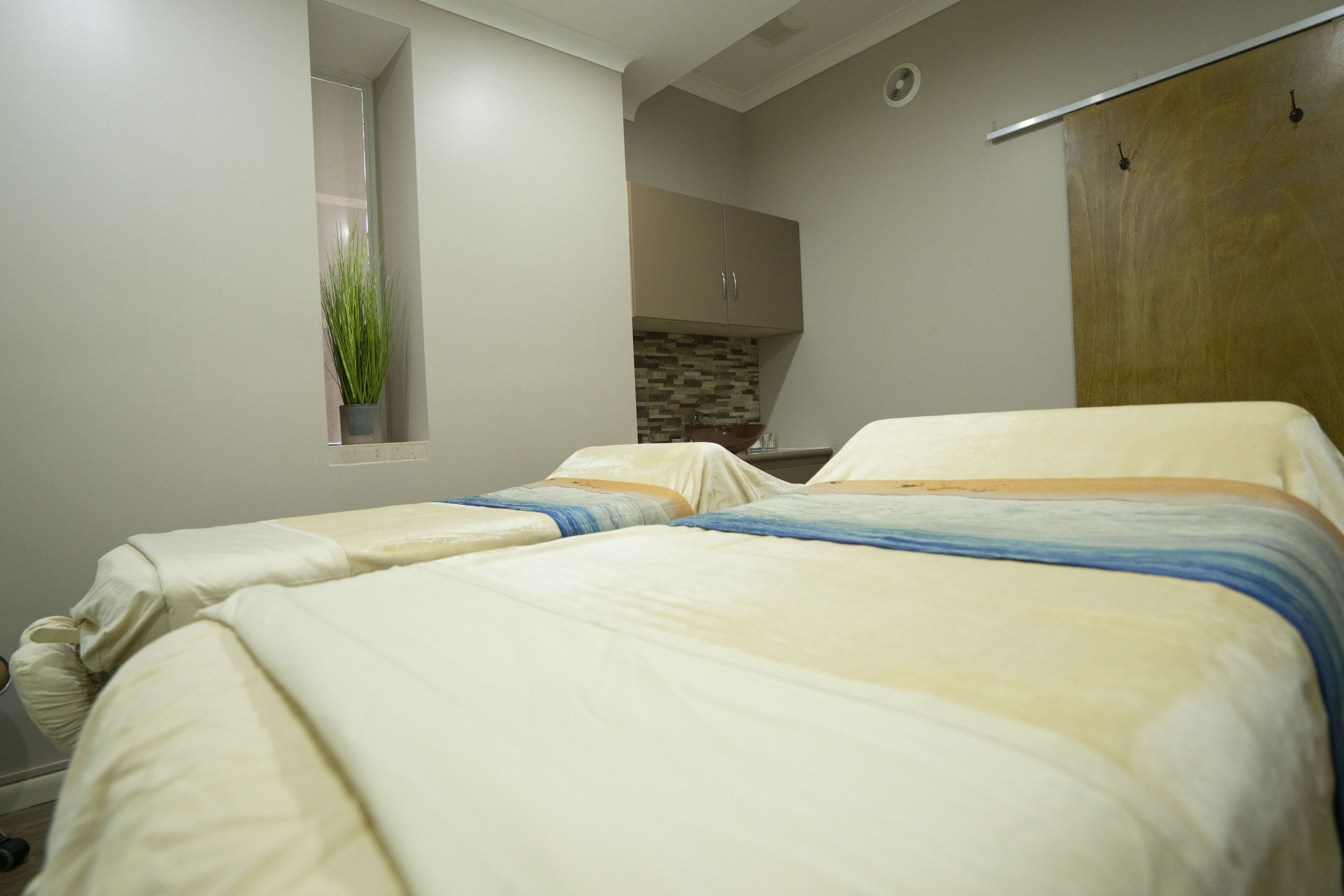 Un dormitorio con dos camas, una ventana pequeña con una planta, una kitchenette y un armario de madera.