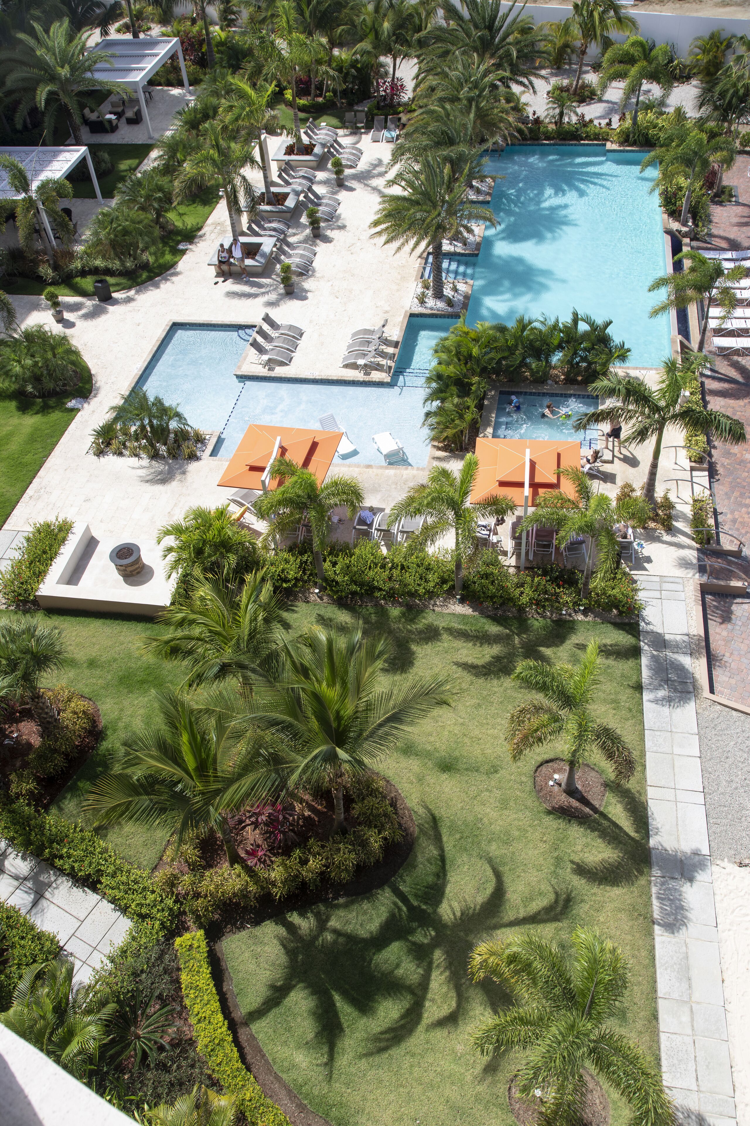 Luchtbeeld van een resort met twee zwembaden, ligbedden, palmbomen en een verzorgd gazon.