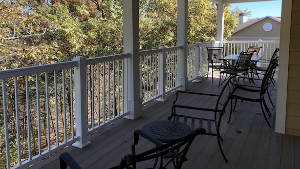 A covered outdoor deck with white railings, a table with chairs, and a view of trees and a house in the background. - 5-Star Branson Hills Villa - 4 Master Suites, Sleeps Up to 10, Pool & Golf (Branson)