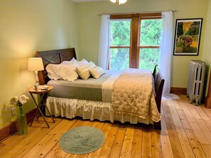 A bedroom with a wooden floor, a bed with a white and green bedspread, a bedside table with a lamp, a radiator, and a poster on the wall.