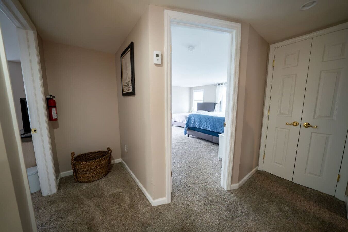 A hallway leading to a bedroom with a bed, a framed picture, a fire extinguisher, and a basket.