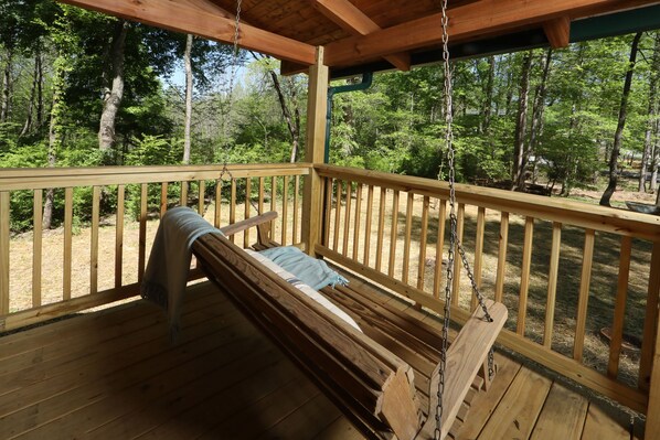 A wooden swing chair on a covered porch with a view of a forest.