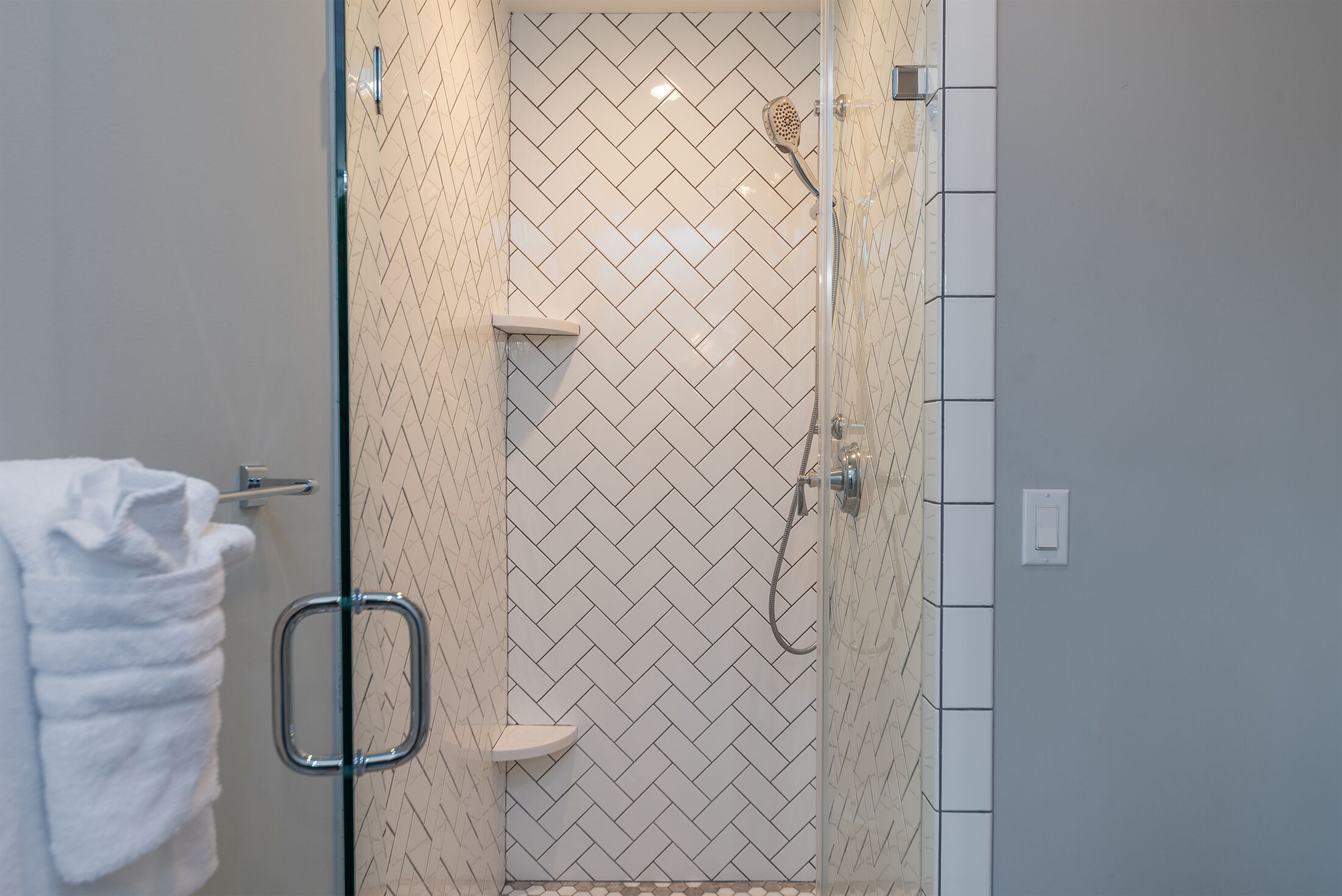 Een douche met glazen wanden, een visgraatmotief van tegels, een witte handdoek hangend aan de deur en een klein plankje.