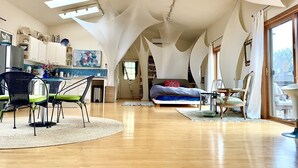 Um quarto amplo no estilo loft, com uma cama grande, área de jantar com mesa e cadeiras, e cozinha com armários brancos e painel de fundo azul.