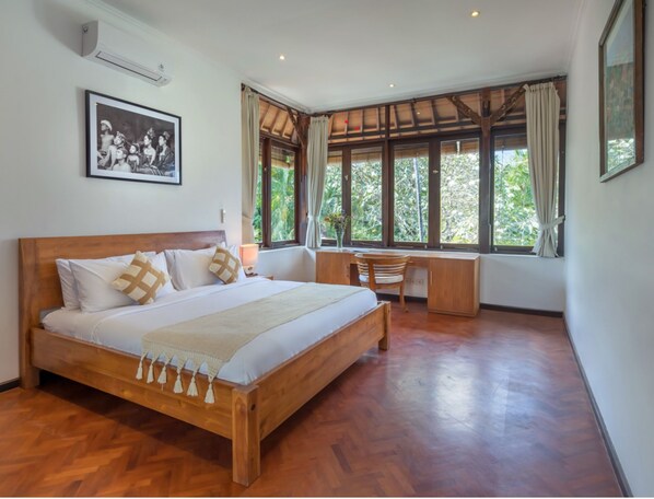 A spacious bedroom with a large bed, a wooden desk, and a view of lush greenery through large windows.