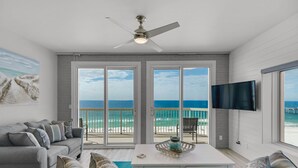 A living room with a large window overlooking a beach, a ceiling fan, and a flat-screen TV mounted on the wall.