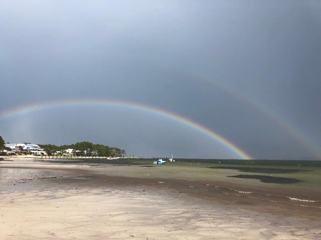 A beach with a double rainbow, calm sea, and houses in the background.