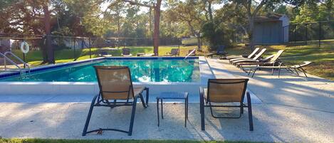 Une piscine avec des chaises longues et une petite table, entourée d’arbres et délimitée par une clôture.