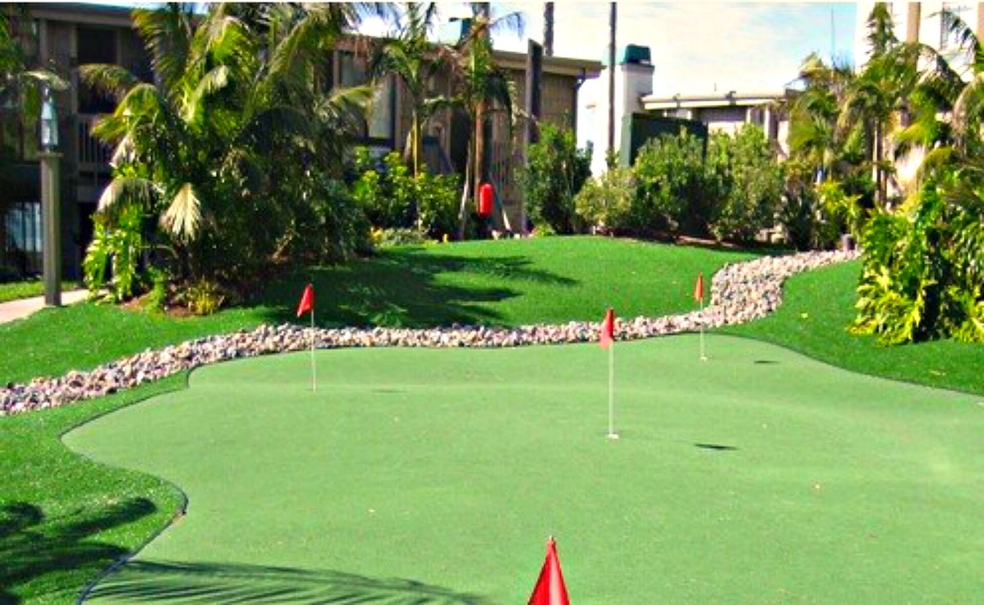 A golf course with a well-maintained green, red flags marking the holes, and a stone wall running along the edge.