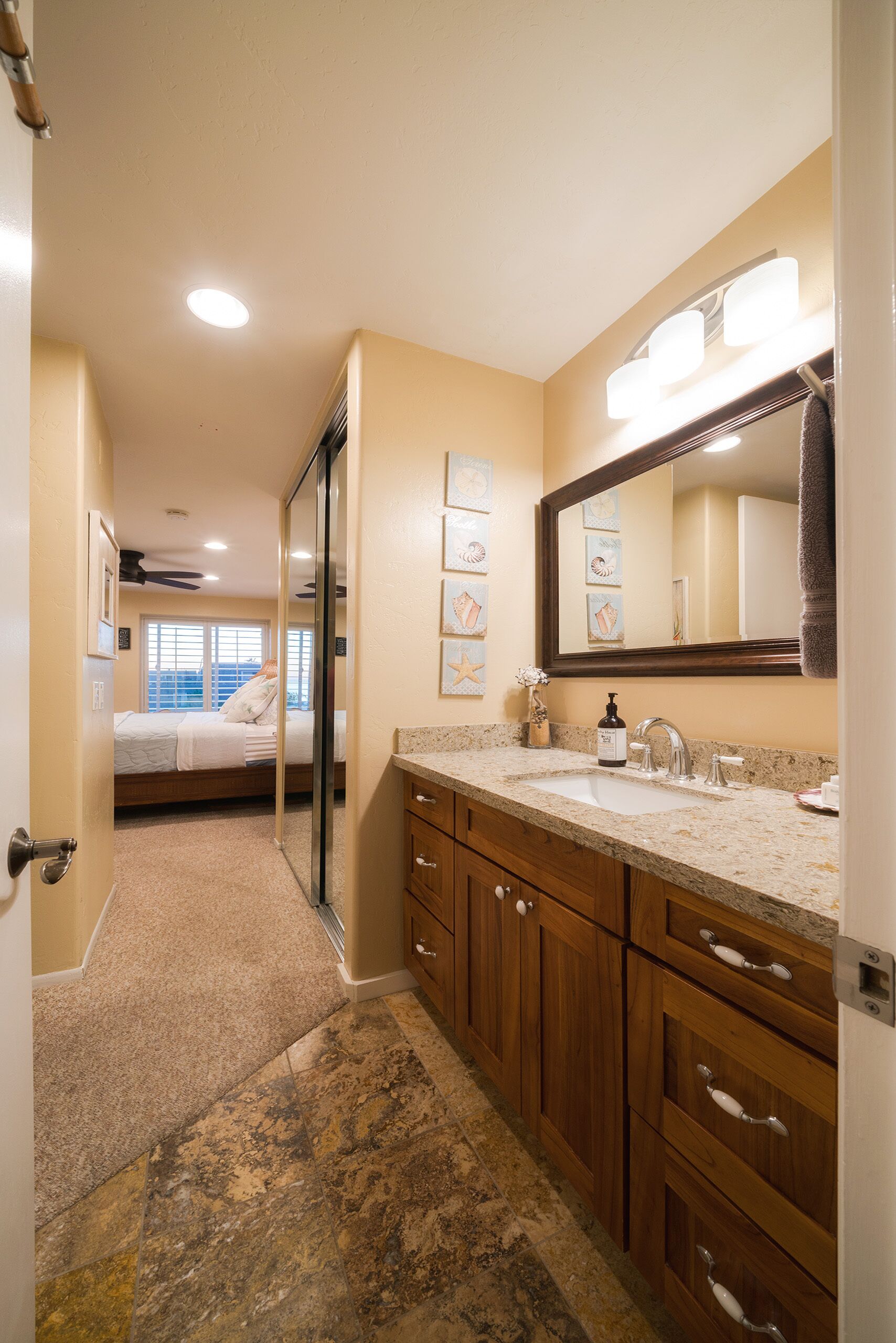 Un baño con un espejo grande, un lavamanos y un mueble de tocador con cajones. Hay una puerta que da a otra habitación.