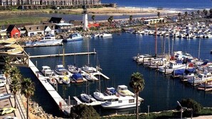 Un puerto deportivo con numerosas embarcaciones amarradas, un muelle con algunos edificios y una playa al fondo.