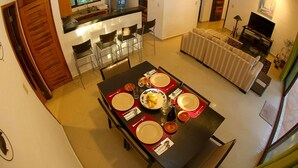 A dining area with a table set for four, a kitchen with modern appliances, a living room with a sofa, and a TV. - 2 Bedroom Apartment In Quiet Secure Residential Complex Surrounded By Nature (Akumal)
