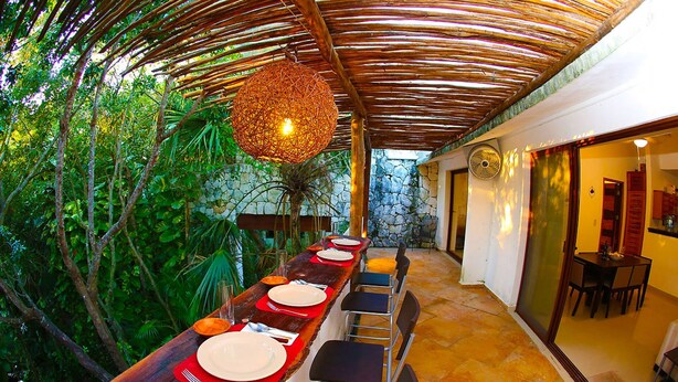 An outdoor dining area with a long wooden table, chairs, and a hanging light fixture. - 2 Bedroom Apartment In Quiet Secure Residential Complex Surrounded By Nature (Akumal)