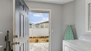 Une buanderie équipée d’un lave-linge, d’un étendoir et d’une porte donnant sur un espace extérieur avec une clôture blanche et un ciel dégagé.