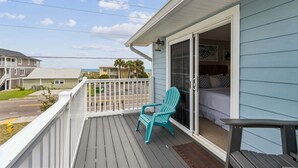 Un balcon avec un fauteuil Adirondack turquoise, un lit avec des oreillers et une vue sur la rue et les autres bâtiments.