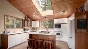 A modern kitchen with a central island, wooden ceiling, large windows, and a skylight.