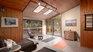 A cozy room with a wooden ceiling, large windows, a sofa, a chair, a small table, and a dresser.