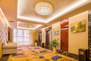 A hotel lobby with a patterned carpet, chandeliers, a sofa, potted plants, and artwork on the walls.