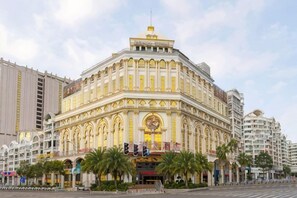 Un edificio grande y ornamentado con un reloj destacado y ventanas arqueadas, rodeado de palmeras y otros edificios.