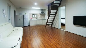 A spacious living area with a white sofa, wooden flooring, a kitchen with white cabinets, a staircase, and a mounted TV. - Jeju J Pension (Seogwipo)