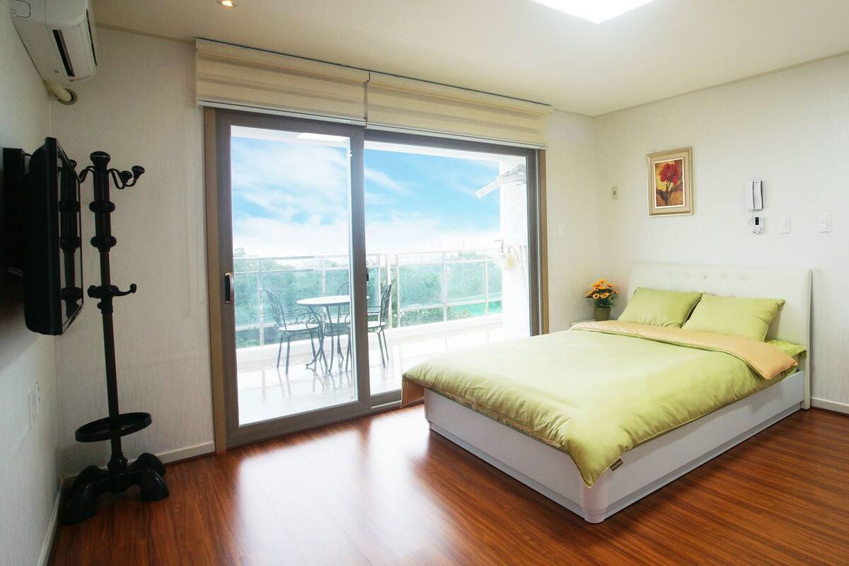A bedroom with a bed, a sliding glass door to a balcony, a small table with a vase of flowers, and a white door.