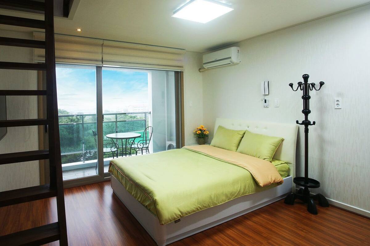 A bedroom with a bed, a standing lamp, a ladder, and a balcony with a table and chairs.