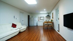 A modern living room with a white sofa, a dining table with chairs, a kitchen area with a stove, and a TV mounted on the wall. - Jeju J Pension (Seogwipo)
