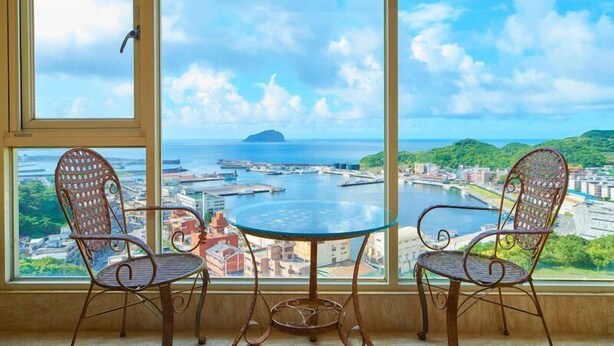 A room with two metal chairs and a glass table, overlooking a harbor with boats and buildings. - HF Keelung 61 (Keelung)