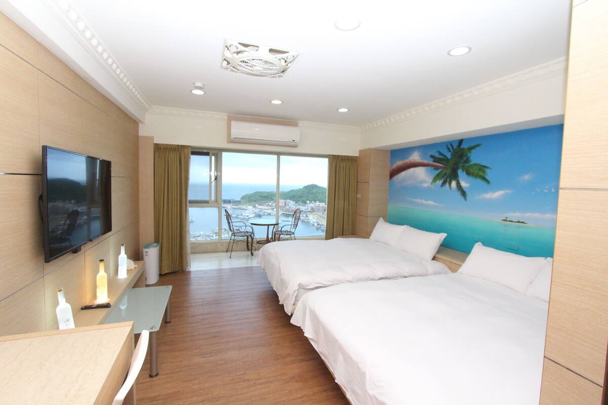 A hotel room with two beds, a TV, a desk with a chair, and a view of the sea and a palm tree through large windows.