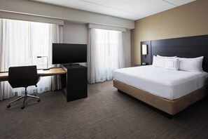 Ein Hotelzimmer mit einem großen Bett, einem Schreibtisch mit Computer, einem Fernseher und einem Fenster mit Vorhängen.