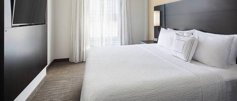 A hotel room with a large bed, a flat-screen TV mounted on the wall, a nightstand with a lamp, and a window with sheer curtains.