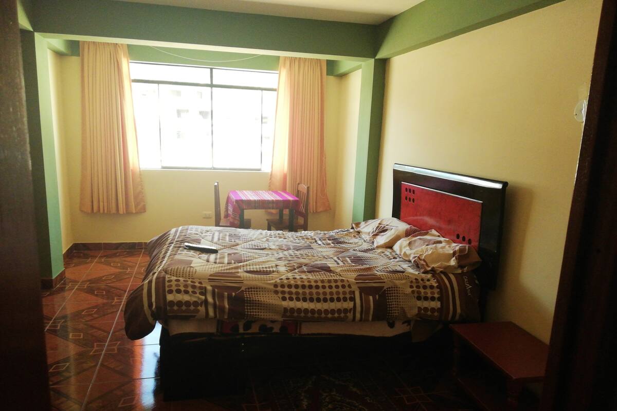 Un dormitorio con cama, una mesita y una silla. Hay una ventana con cortinas y el suelo es de baldosas.